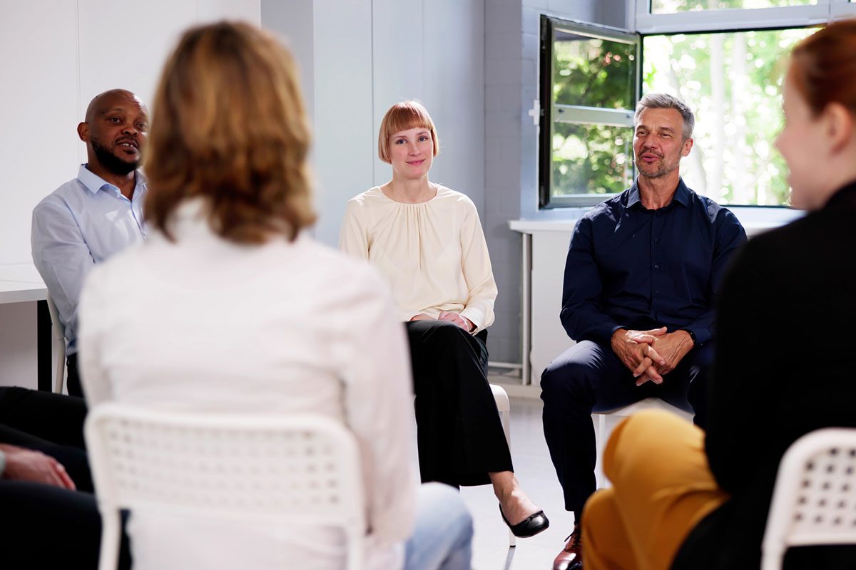 A group of diverse adults sit in a circle in a therapeutic setting for a group therapy session