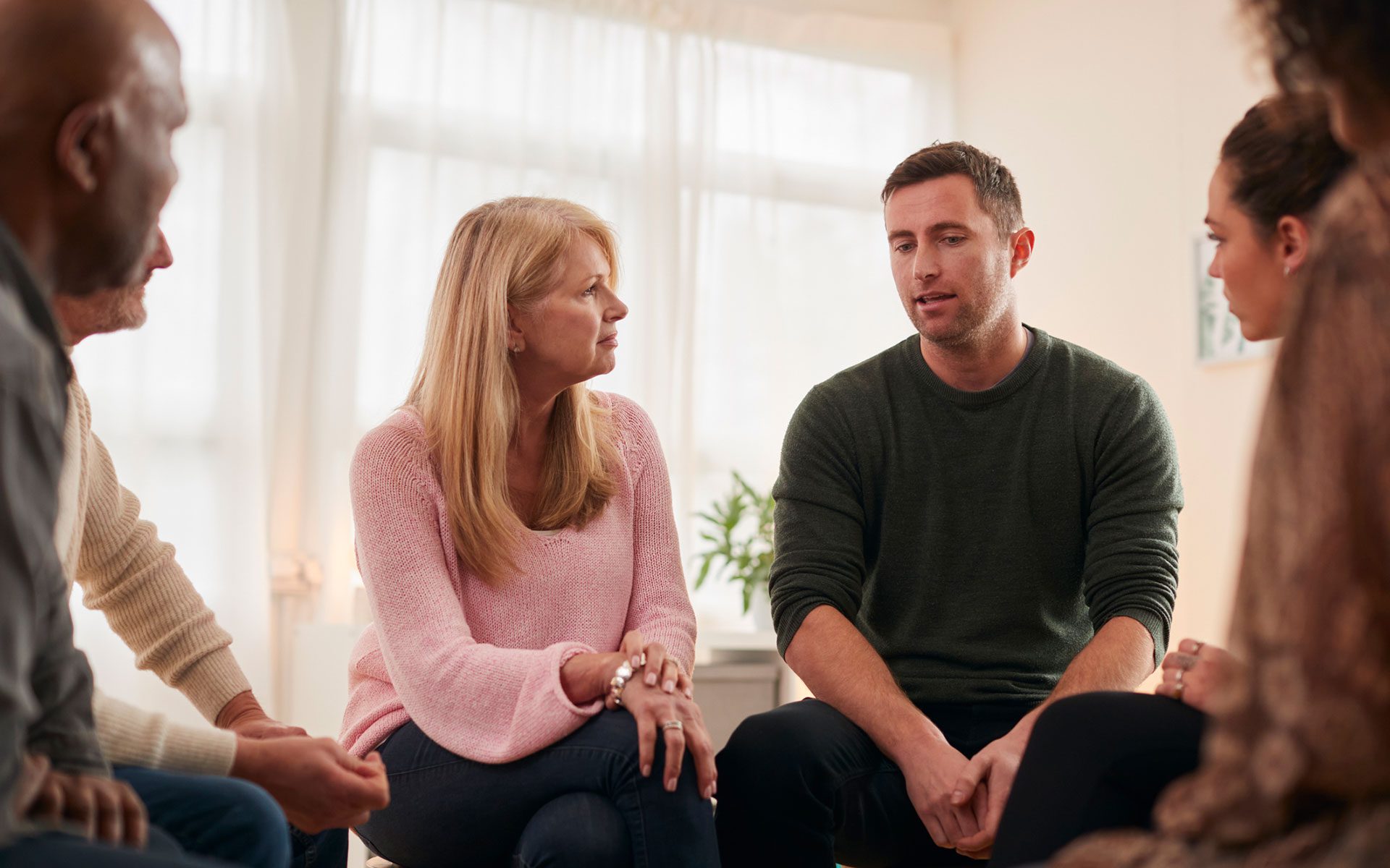 A small group of adults participating in group therapy listen as one participant, a man, speaks