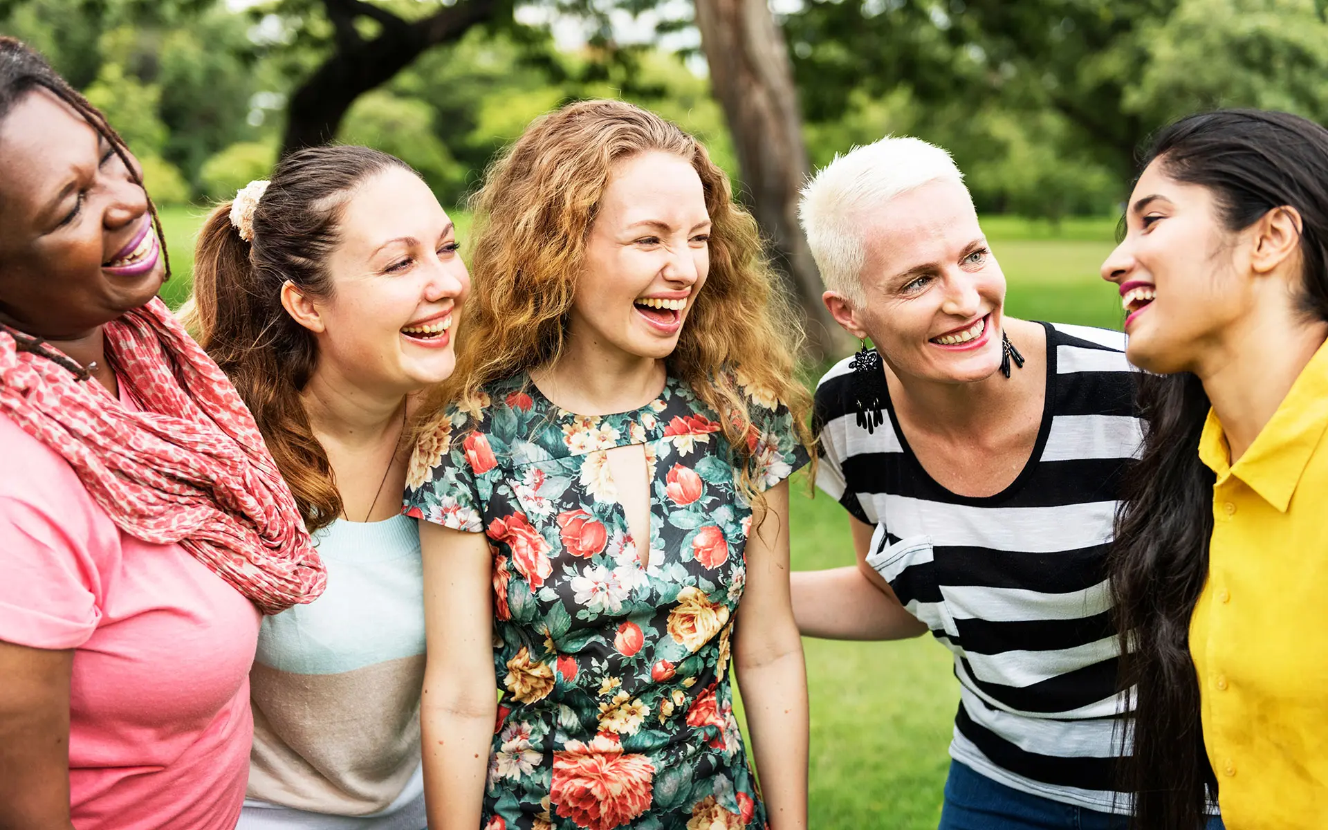 A group of woman laugh and hold each other should-to-shoulder as they stand in the park